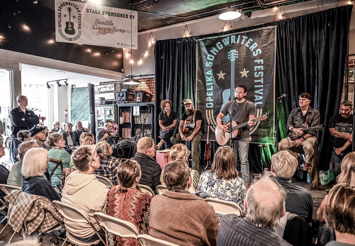 A full room of listeners in folding chairs facing a songwriters round on the OSF stage downtown