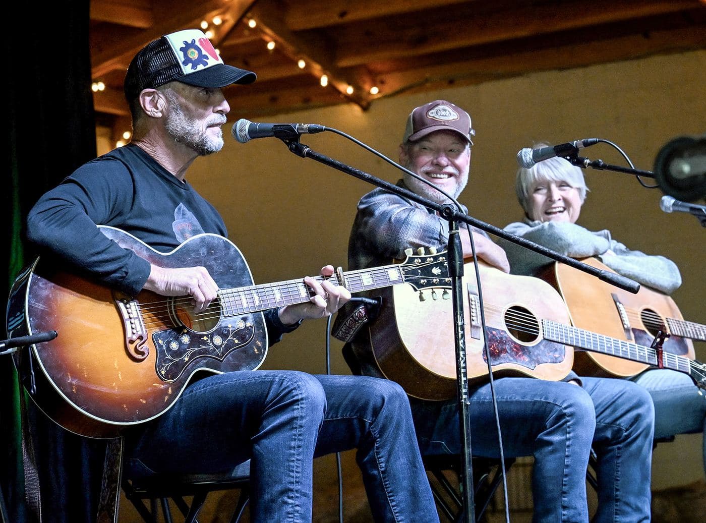 Three songwriters seated in a row playing acoustic guitars under warm string lights