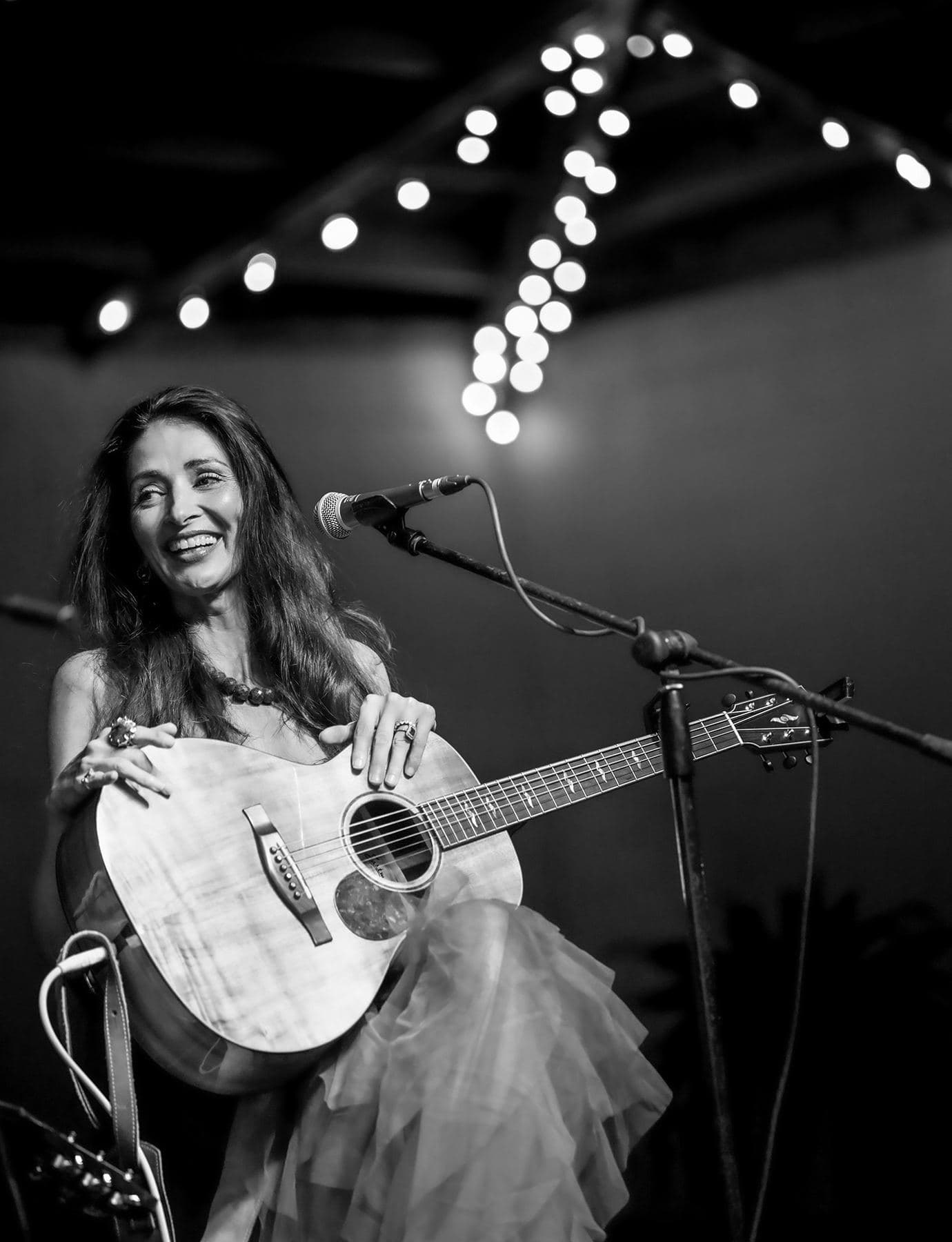 A songwriter smiling mid-song under a string of stage lights, in black and white