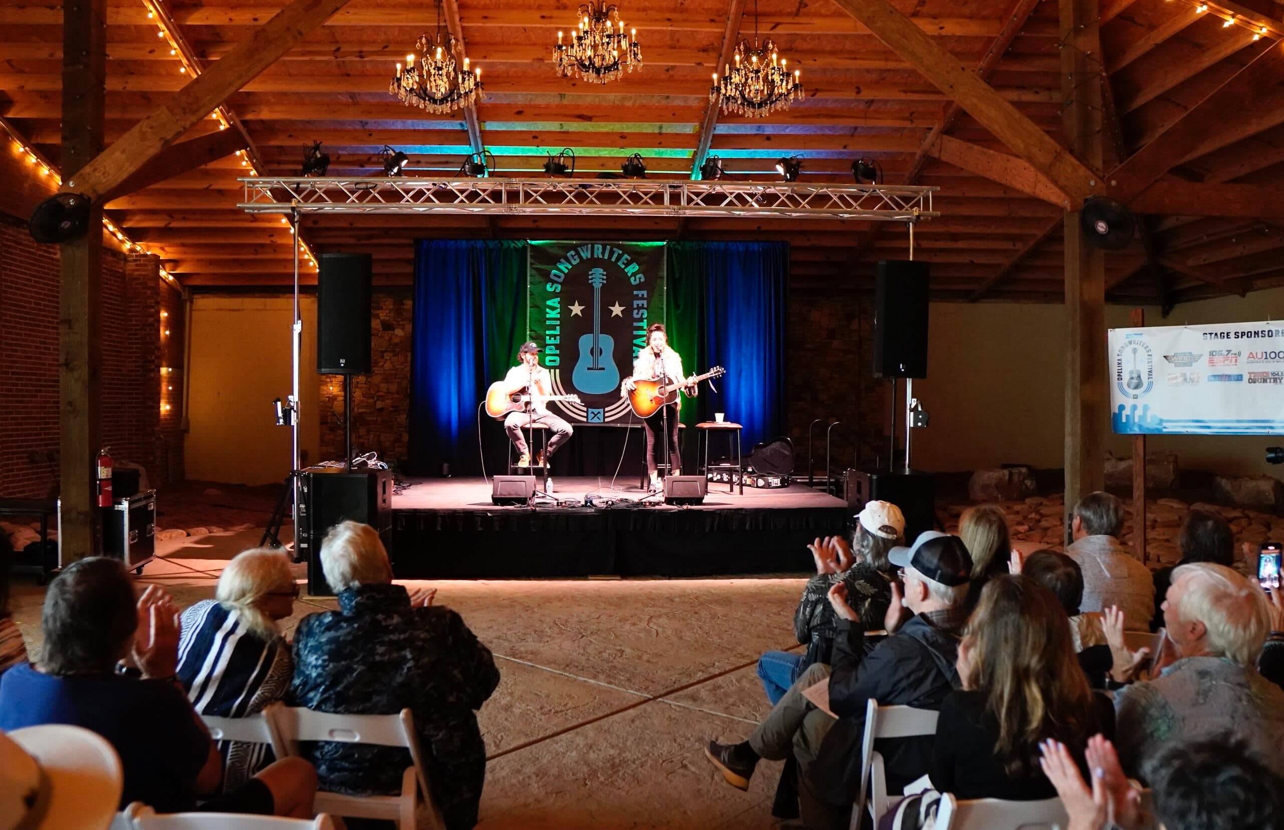 Two songwriters performing on the Bottling Plant Pavilion stage beneath chandeliers